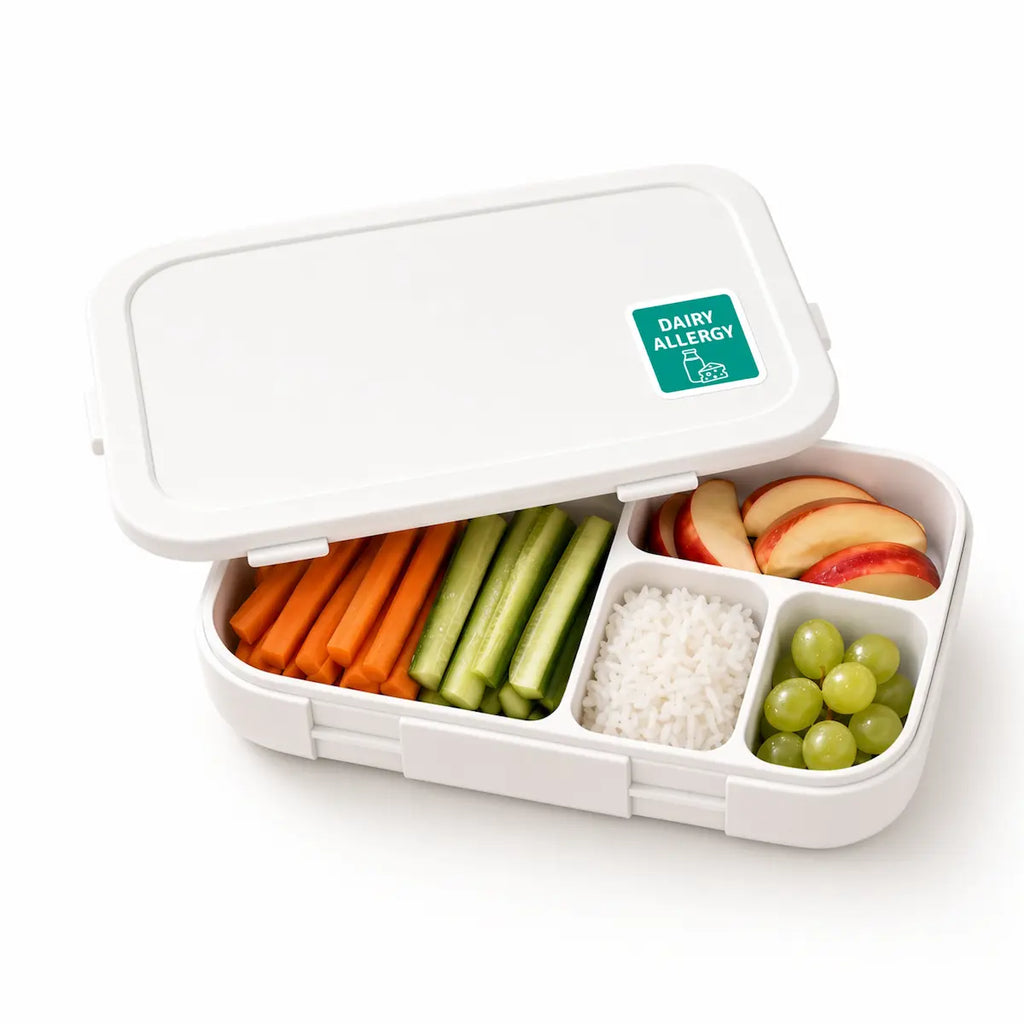 Open white lunch box with a green dairy allergy warning sticker on the lid, containing a sandwich, apple slices, crackers, and green grapes