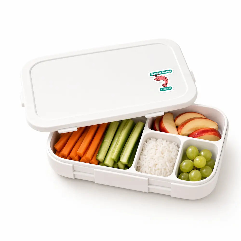 A cartoon shrimp shellfish allergy alert sticker on a white bento lunchbox packed with carrots, cucumbers, rice, apple slices, and grapes for school meal identification.