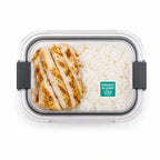 Clear plastic lunch box with a green tree nut allergy warning sticker on the lid, containing sliced grilled chicken and white rice.