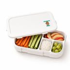 A cartoon tree nut allergy alert sticker on a white bento lunchbox filled with carrots, cucumbers, rice, apple slices, and grapes for daycare and school allergy awareness.