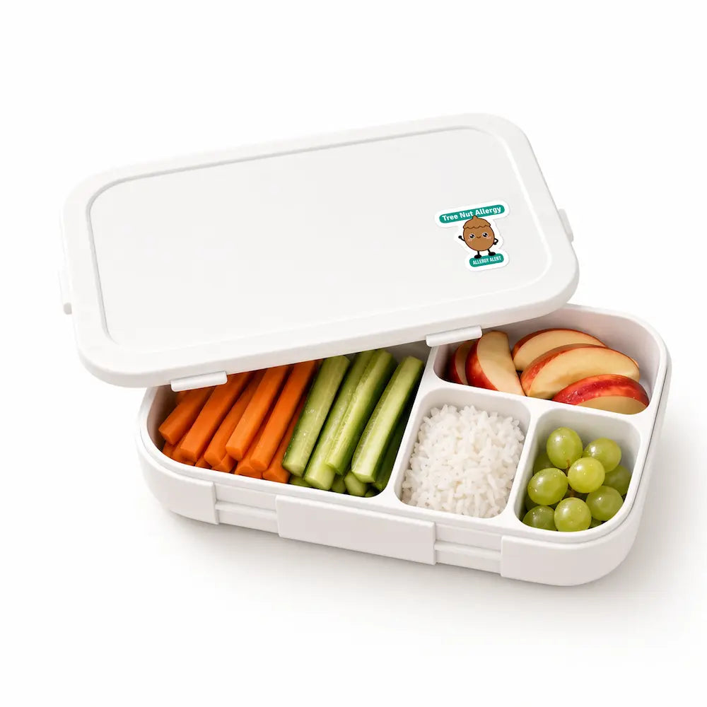 A cartoon tree nut allergy alert sticker on a white bento lunchbox filled with carrots, cucumbers, rice, apple slices, and grapes for daycare and school allergy awareness.